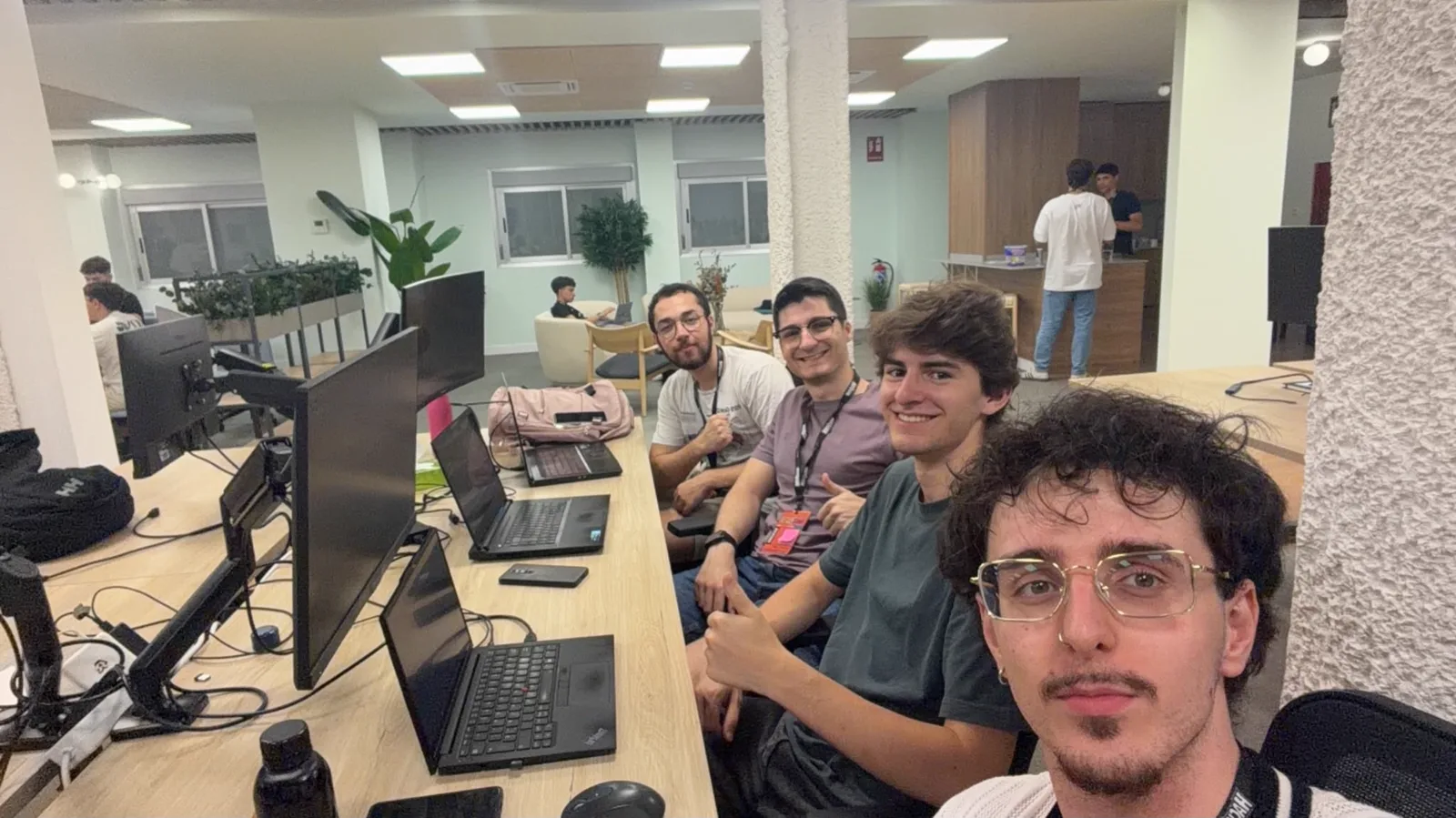 Selfie de cuatro integrantes de KermitPanic sonriendo en una mesa larga con portátiles y monitores, en una oficina amplia con plantas y otros participantes al fondo.