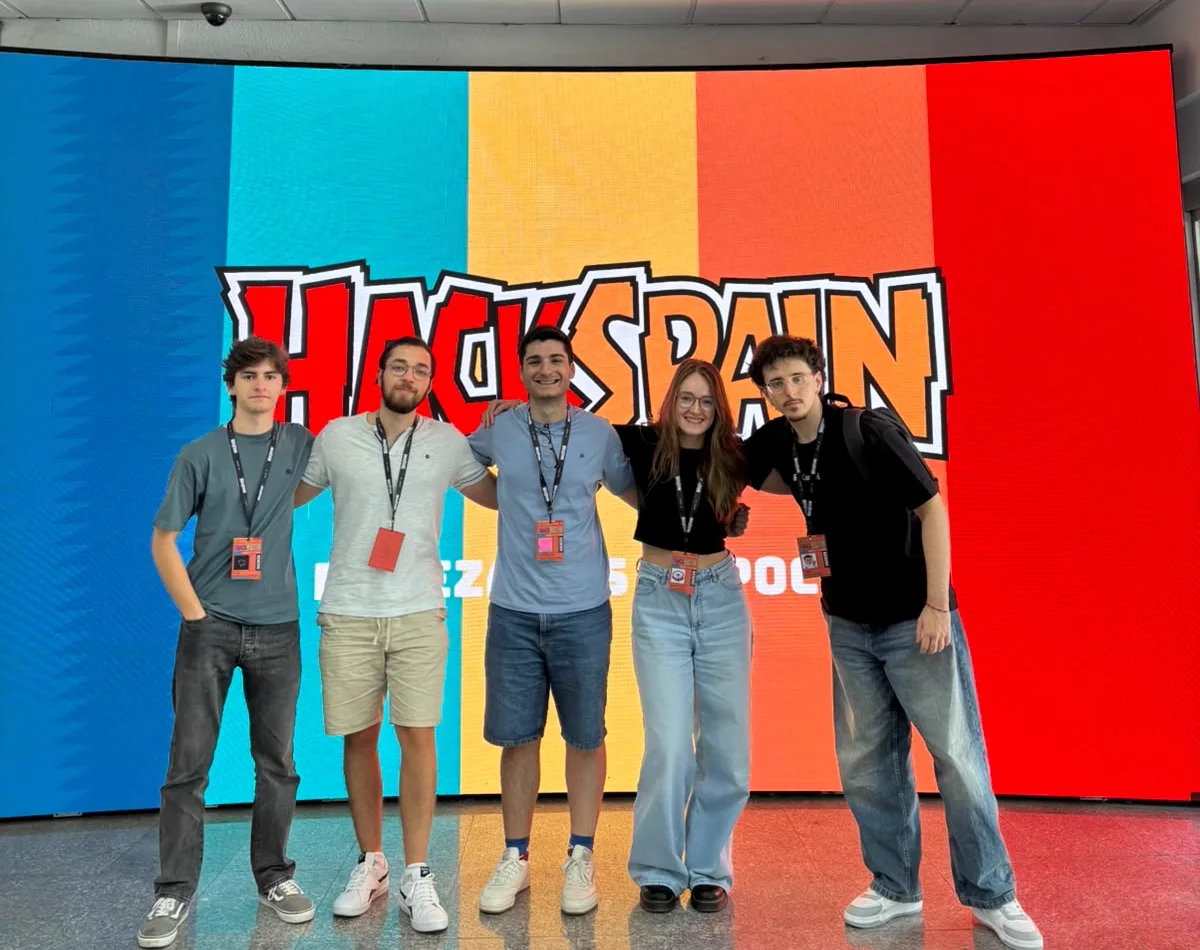 Las cinco personas de KermitPanic, con sus acreditaciones al cuello, posan abrazadas delante de una gran pantalla con el logo de HackSpain sobre franjas azul, turquesa, amarilla, naranja y roja.